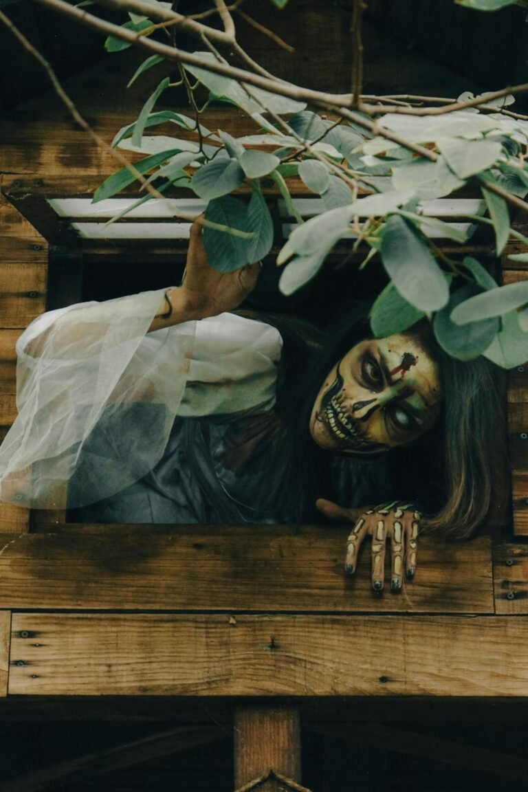 A spooky zombie with face paint reaching out from a window surrounded by leaves.